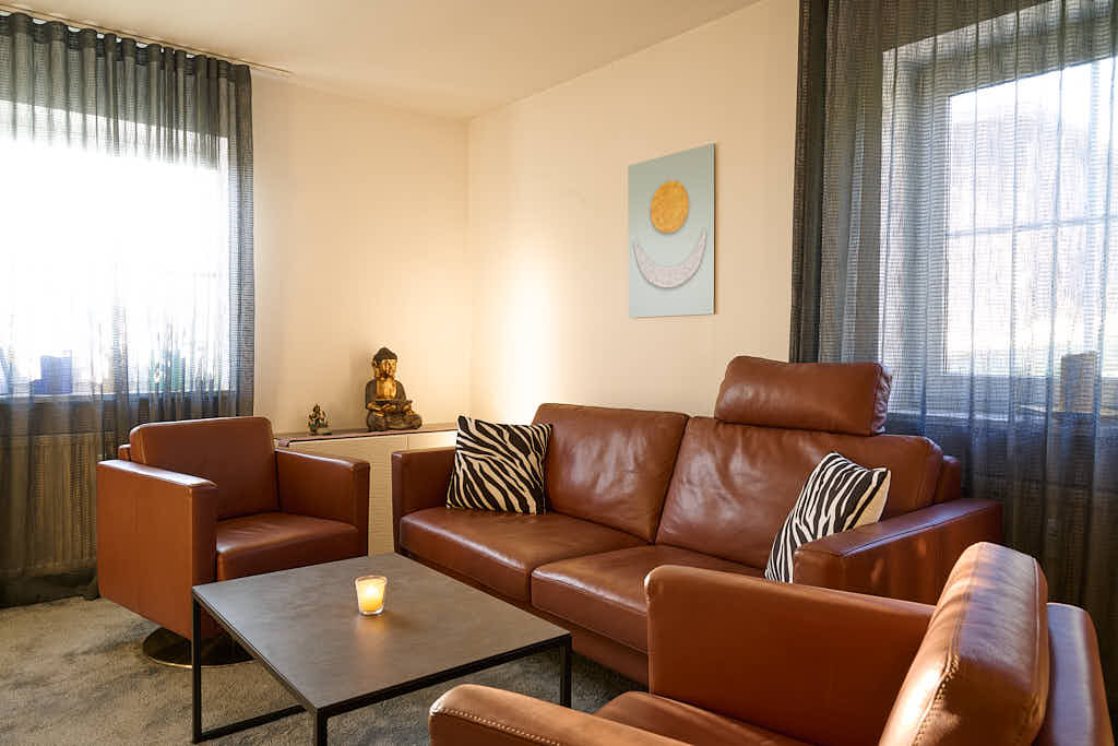 Ein gemütlicher Gesprächsraum mit einem braunen Ledersofa und zwei passenden Sesseln, einem schwarzen Couchtisch mit einer brennenden Kerze, Kissen mit Zebramuster, durchsichtigen Vorhängen und einer Buddha-Statue auf einem Beistelltisch. An der Wand hängt ein Gemälde.