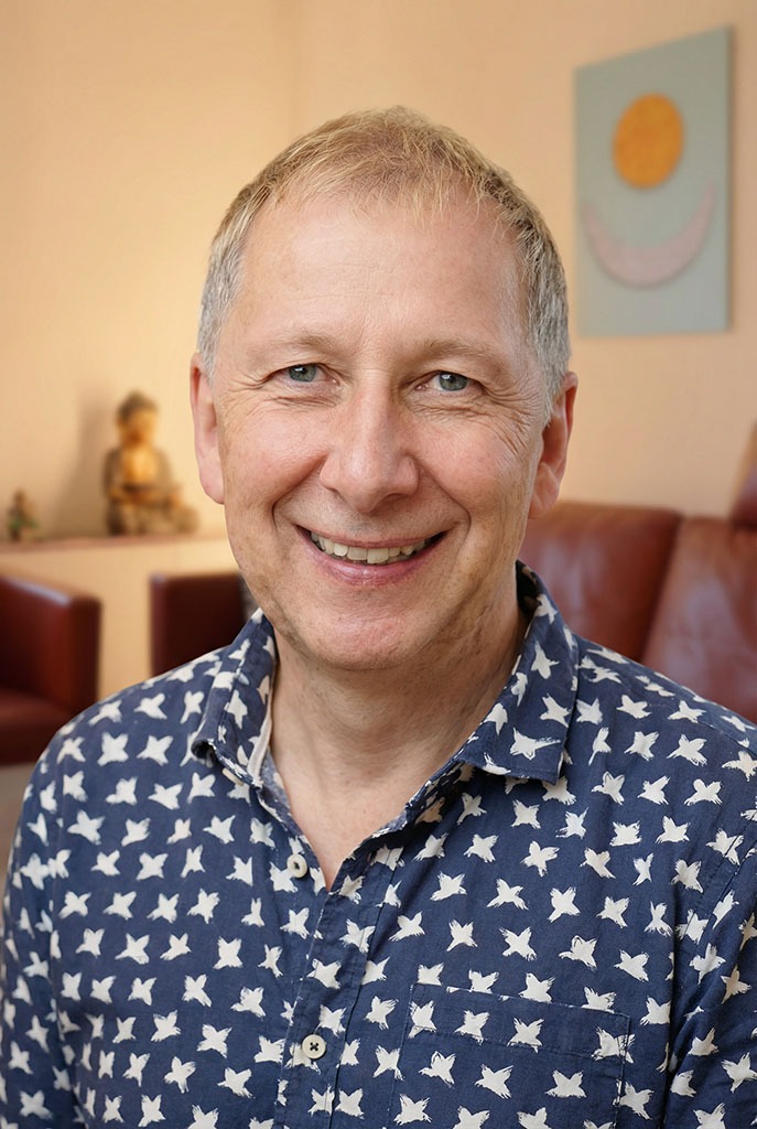 Ein lächelnder Coach mittleren Alters mit kurzen blonden Haaren trägt ein blaues Hemd mit einem weißen Sternenmuster. Er sitzt drinnen in einem warm beleuchteten Raum mit roten Stühlen und einem Meditationsbild hinter ihm.